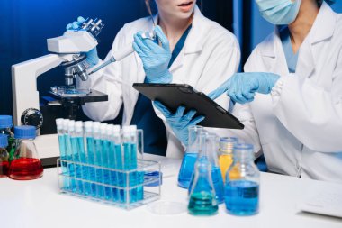 Bir tıp laboratuarında araştırma yapan genç bilim adamları, ön plandaki bir araştırmacı, tıp laboratuvarında mikroskop ve dijital tablet kullanıyor.  