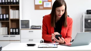 Genç ve güzel bir kadın iş yerinde otururken akıllı telefon ve dizüstü bilgisayar kullanıyor. Modern ofiste işe odaklanmış. 