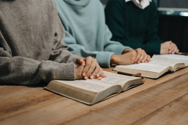 Group of Christians sit together and pray around a wooden table with blurred open Bible pages in their homeroom. Prayers for brothers, faith, hope, and seek the blessings of God. clipart