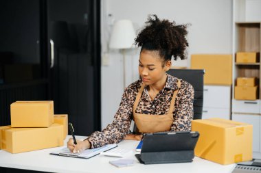 Afro-Amerikalı girişimci kadın çevrimiçi alışveriş siparişleri için paket ürün kutuları hazırlıyor. Başlangıçta küçük işletme KOBİ kavramı.