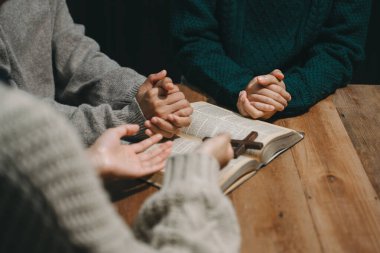 Group of Christians sit together and pray around a wooden table with blurred open Bible pages in their homeroom. Prayers for brothers, faith, hope, and seek the blessings of God. clipart