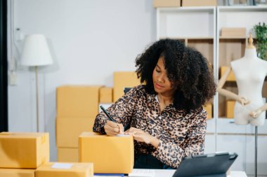 Afro-Amerikalı girişimci kadın çevrimiçi alışveriş siparişleri için paket ürün kutuları hazırlıyor. Başlangıçta küçük işletme KOBİ kavramı.