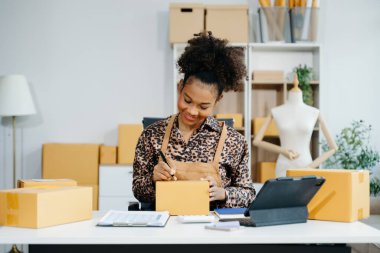 Afro-Amerikalı girişimci kadın çevrimiçi alışveriş siparişleri için paket ürün kutuları hazırlıyor. Başlangıçta küçük işletme KOBİ kavramı.