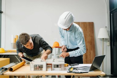 Mimaride profesyonel işbirliği. İki mühendis, dizüstü bilgisayarlar, tabletler ve ölçekli modeller gibi araçlar kullanarak inşaat planları üzerinde çalışıyor..