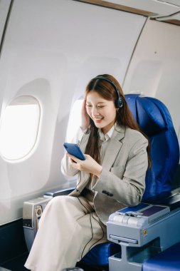 Uçakta birinci sınıftaki genç Asyalı iş kadını kulaklıkla müzik dinlemek için akıllı telefon kullanıyor.  