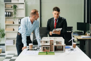 Masanın yanında duran çeşitli iş adamları konut projesinin modeline bakıyor ve dijital tablet kullanıyor. Yeşil iş şirketi ve Güneş Enerjisi Çevre Kenti Kavramı.