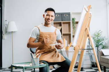 Asyalı erkek ressam resim atölyesinde resim malzemeleri, yağlı boya tabloları, iki tuval sehpası, resim stüdyosunda veya oturma odasında fırçalı yaratıcı bir alan ile sanat çalışması yapıyor..