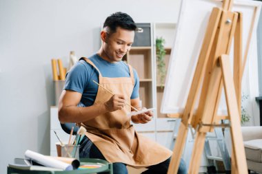 Asyalı erkek ressam resim atölyesinde resim malzemeleri, yağlı boya tabloları, iki tuval sehpası, resim stüdyosunda veya oturma odasında fırçalı yaratıcı bir alan ile sanat çalışması yapıyor..
