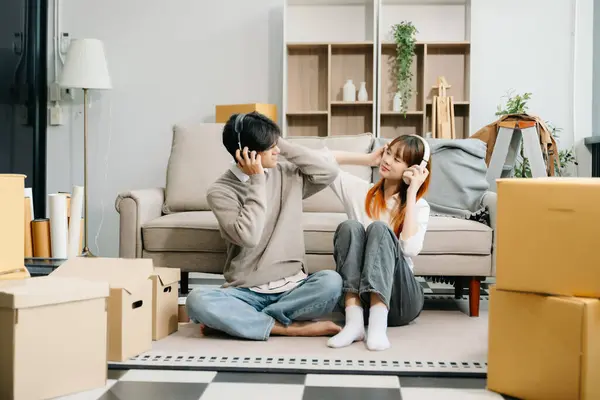 stock image Happy asian young attractive couple man and woman with big boxes moving into a new house, relaxing on the sofa  and listening music in headphones 