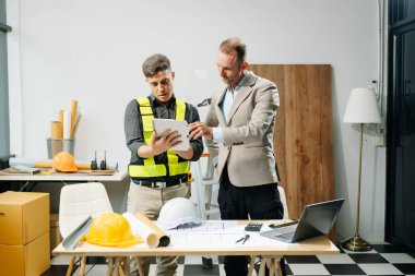 Müteahhit ve mimar planı tablet, dizüstü bilgisayar kullanarak ve büyük bir proje üzerinde çalışmakla ilgili konuşarak ele alıyorlar. İnşaat sürecinde bina içi