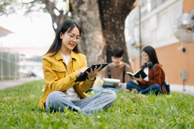 Asyalı öğrenciler kampüs parkında ders çalışıyorlar. Gençler birlikte çimenlerde oturup kitap okuyor ve Green Park 'taki arkadaşlarının önünde tablet kullanıyor.