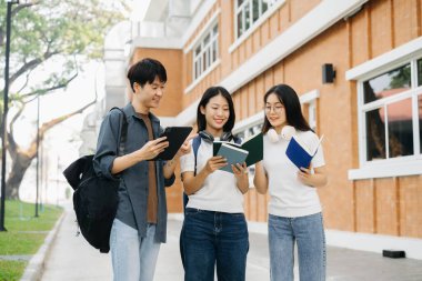 Genç Asyalı üniversite öğrencileri sabahleyin kampüs parkında yan yana duruyorlar. Kadınlar konuşurken adam tablet kullanıyor.
