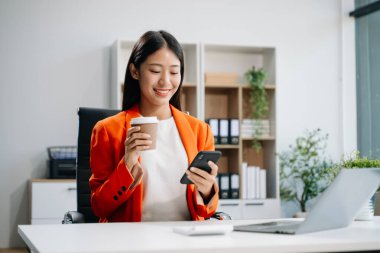 Genç ve güzel bir iş kadını iş yerinde otururken akıllı telefon kullanıyor ve kahve içiyor. İşine odaklan..   