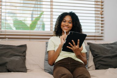 Genç kıvırcık Afrikalı kadın rahat bir evde kanepede tablet kullanıyor.