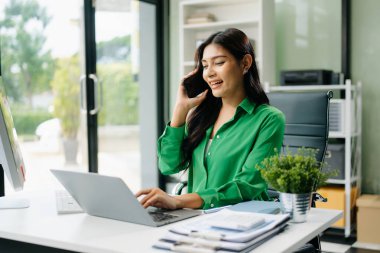 Genç ve çekici Asyalı ofis çalışanı takım elbiseli Laptop bilgisayarında çalışıyor ve akıllı telefondan konuşuyor. 