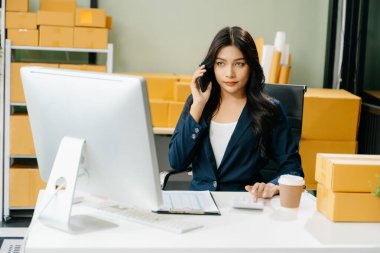 Genç işletme sahibi bilgisayardaki müşterilere internet siparişlerini kontrol ediyor ve akıllı telefondan konuşuyor. Çevrimiçi Alışveriş kavramı.