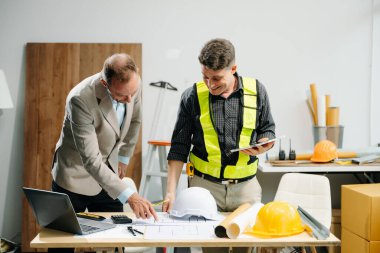 Müteahhit ve mimar, dizüstü bilgisayar ve dijital tablet kullanarak büyük bir proje üzerinde çalışıp çalışmaktan bahsediyorlar.