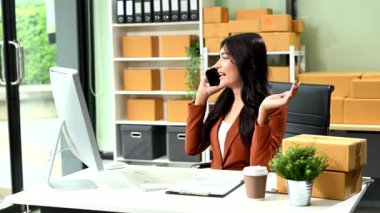 Ofisteki kutuları olan genç bir iş kadını ofisteki internet mağazasında bilgisayarla çalışıyor ve akıllı telefondan konuşuyor. 