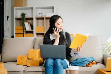  Çevrimiçi satışları yöneten genç bir iş kadını ve modern bir ev ofisinden gelen nakliyat, dizüstü bilgisayar kullanarak ve elinde not defteriyle akıllı telefonla konuşarak.  