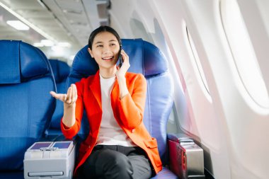 Asyalı bir kadın uçakta bir koltukta oturuyor ve seyahat konseptinde akıllı telefondan konuşuyor..
