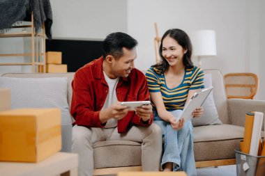 Asyalı genç ve çekici bir çift dijital tablet ve akıllı telefonu ev dekorasyonu için online alışveriş mobilyaları olarak kullanıyor.