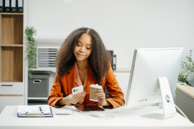 Akıllı telefon kullanan ve modern ofiste kahve içen kendine güvenen Asyalı iş kadını.