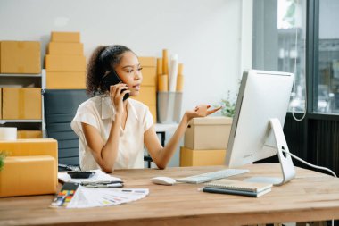 Başlangıçta küçük işletme KOBİ 'si, akıllı telefondan konuşan girişimci kadın ve yeni siparişleri kontrol etmek için bilgisayarı kullanmak, online alışveriş kavramı, karton kutular arka planı