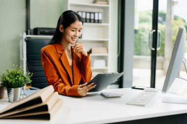 Asyalı bir kadın masada oturuyor ve ofiste bilgisayar kullanıyor, panoya not alıyor. Teknolojiyle Mali ve Pazarlama Yönlendirme