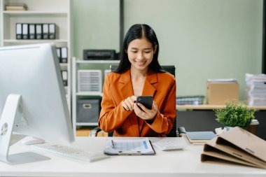 Masada oturan Asyalı kadın ofiste bilgisayar ve akıllı telefon kullanıyor. Teknolojiyle Mali ve Pazarlama Yönlendirme