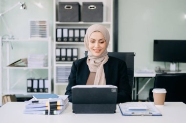 Tabletle çalışan, büro masasında telefonla konuşan tesettürlü genç Arap bayan girişimci. 