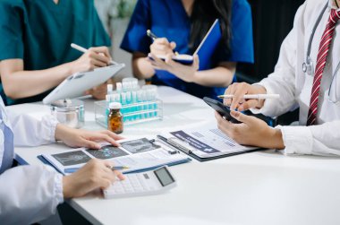 Tıbbi ekip beyaz önlüklü ve ameliyat önlüklü doktorlarla bir toplantı yapıyor. Masada oturmuş dijital tablet kullanan bir hastanın teşhisini tartışıyorlar. 