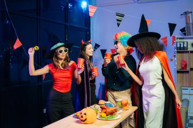 Cadılar Bayramı partisi için kostüm giymiş genç iş arkadaşları içki içip ofiste kahkaha atıyorlar. Ekip çalışması ve şenlik ruhu.