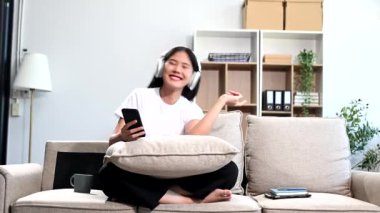 Genç Asyalı kadın evde kanepede otururken kulaklığı ve akıllı telefonuyla müzik dinliyor.