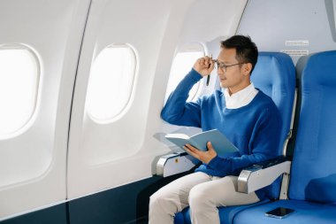 Asyalı bir adam uçak koltuğunda oturuyor, seyahate çıkarken gündemine notlar yazıyor.. 