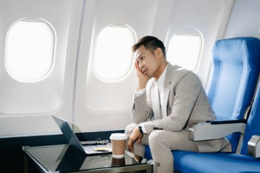 Üzgün Asyalı iş adamı bir uçak koltuğunda oturuyor ve laptopla çalışıyor. Seyahate çıkıyor..