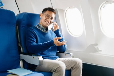 Asyalı bir adam uçakta bir koltukta oturuyor ve akıllı telefondan konuşuyor.