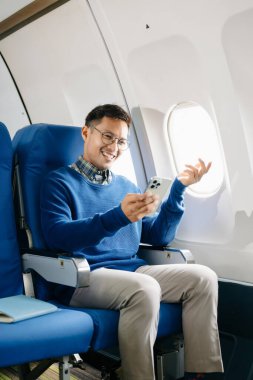 Asyalı bir adam uçağın koltuğunda oturuyor. Tatile gidiyor, akıllı telefon kullanıyor.. 