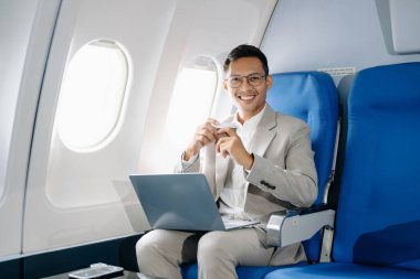 Asyalı bir adam uçakta oturmuş dizüstü bilgisayar kullanıyor ve kahve içiyor.
