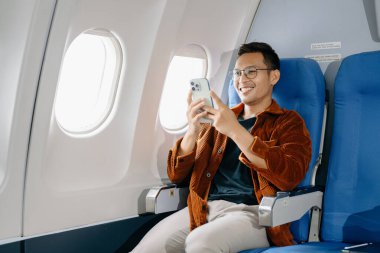 Asyalı adam uçakta bir koltukta oturuyor ve akıllı telefon kullanıyor.