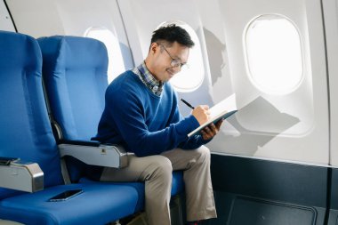Asyalı bir adam uçak koltuğunda oturuyor, seyahate çıkarken gündemine notlar yazıyor.. 