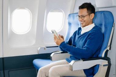 Asyalı adam uçakta bir koltukta oturuyor ve akıllı telefon kullanıyor.