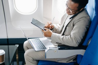 Gülümseyen Asyalı işadamı bir uçak koltuğunda oturuyor, telefonla konuşuyor, laptopta ve tablette çalışıyor ve seyahate çıkıyor..