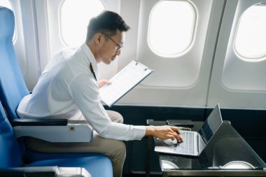 Yolcu koltuğunda üniformalı Asyalı erkek pilot uçuş planını kontrol ediyor ve dizüstü bilgisayar kullanıyor. 