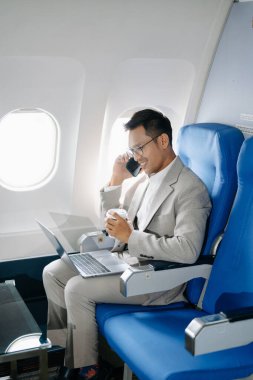 Asyalı bir adam uçakta bir koltukta oturup telefonla konuşurken dizüstü bilgisayar kullanıyor ve kahve içiyor.