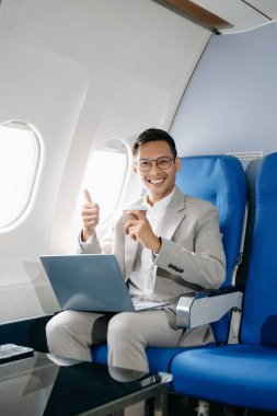 Asyalı bir adam uçakta oturmuş dizüstü bilgisayar kullanıyor ve kahve içiyor.