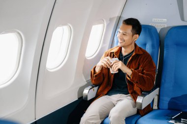 Mutlu Asyalı bir adam uçağın koltuğunda oturmuş tatile çıkıyor, kahve içiyor ve kulaklıkla müzik dinliyor..