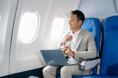 Asyalı bir adam uçakta oturmuş dizüstü bilgisayar kullanıyor ve kahve içiyor.