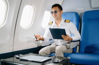 Üniformalı ve güneş gözlüklü gülümseyen Asyalı erkek pilot yolcu koltuğunda uçuş planını kontrol ediyor.