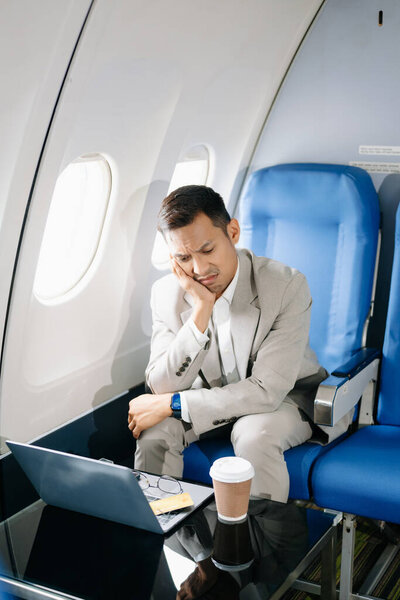 грустный азиатский бизнесмен, сидящий в кресле самолета и работающий на ноутбуке собирается в отпуск.