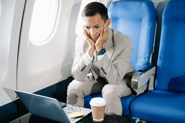 грустный азиатский бизнесмен, сидящий в кресле самолета и работающий на ноутбуке собирается в отпуск.
